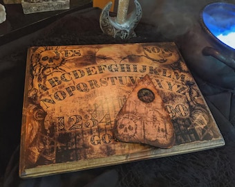 May include: A vintage-style Ouija board with a planchette. The board is made of wood with a distressed finish, featuring letters, numbers, and the words "YES" and "NO." The planchette is also wooden, with a circular viewing hole and skull details.