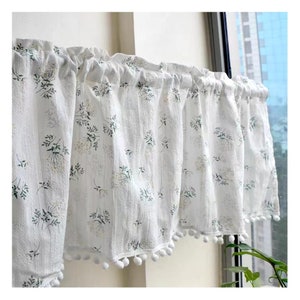 Puede incluir: Cenefa floral blanca con un delicado estampado de flores verdes y amarillas. La tela tiene un aspecto texturizado y está adornada con pequeños pompones blancos en el borde inferior. La cenefa cuelga frente a una ventana.
