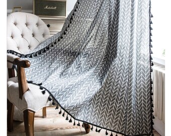 May include: A white and gray chevron patterned curtain with black tassel trim. The curtain is hanging in a room with a chair and a speaker.