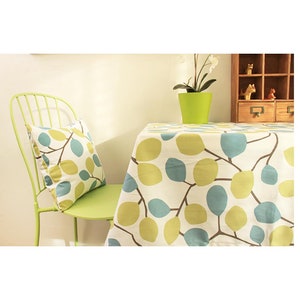 May include: A green metal chair with a leaf-patterned pillow next to a table covered with a matching tablecloth. The tablecloth and pillow feature a white background with blue and green leaf designs. A green pot with a plant sits on the table.