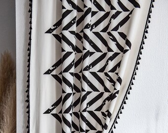 May include: A white curtain with a bold black geometric pattern of diagonal stripes and triangles. The edges are trimmed with black tassels. The curtain is hanging in front of a white wall.