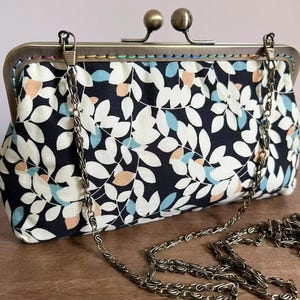 Puede incluir: Un bolso de mano tipo clutch con estampado floral, cierre metálico y correa de cadena. La tela presenta un patrón de hojas blancas con detalles en azul y melocotón sobre un fondo azul marino. El cierre y la cadena son de color bronce.