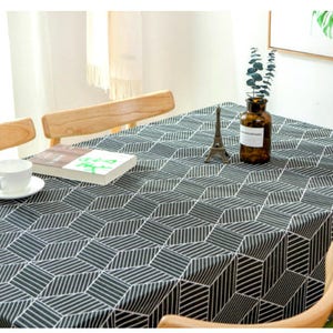 Könnte beinhalten: Eine geometrisch gemusterte Tischdecke in Schwarzweiß mit modernem Design. Die Tischdecke liegt auf einem Holztisch mit Stühlen und einer Vase mit Grünpflanzen.