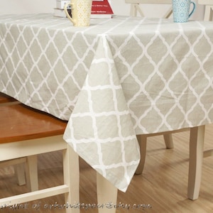 Op de afbeelding: Een beige tafelkleed met een wit geometrisch patroon. Het tafelkleed is over een houten tafel gelegd met twee witte stoelen.