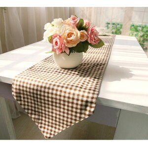 Puede incluir: Un camino de mesa a cuadros marrones y blancos con un jarrón de cerámica blanco lleno de flores rosas y blancas.