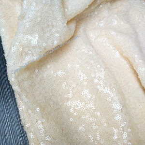 Op de afbeelding: Close-up van ivoor kleurige stof met kleine, witte pailletten. De stof is gevouwen en heeft een textuur.