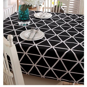 May include: A black and white geometric patterned tablecloth covers a table set for a meal. White plates, silverware, and a turquoise vase with flowers are visible. The tablecloth's design features white lines forming triangles on a black background.