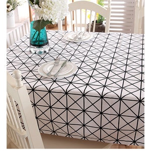 May include: White tablecloth featuring a black geometric design, set with white plates, cutlery, and a turquoise vase holding white flowers. The table is set for a meal, with a white chair in the foreground.