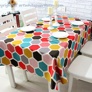 Könnte beinhalten: Eine farbenfrohe geometrische Tischdecke mit einem sechseckigen Muster in Rot-, Gelb-, Blau-, Rosa- und Grautönen. Die Tischdecke liegt auf einem weißen Tisch mit weißen Stühlen.