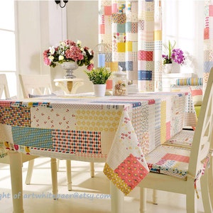 K&ouml;nnte beinhalten: Eine farbenfrohe Patchwork-Tischdecke mit roten, gelben, blauen und wei&szlig;en Quadraten. Die Tischdecke liegt auf einem wei&szlig;en Tisch mit vier wei&szlig;en St&uuml;hlen. Die St&uuml;hle haben Kissen mit einem &auml;hnlichen Patchwork-Design.