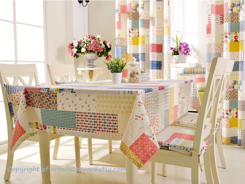 K&ouml;nnte beinhalten: Eine farbenfrohe Patchwork-Tischdecke mit roten, gelben, blauen und wei&szlig;en Quadraten. Die Tischdecke liegt auf einem wei&szlig;en Tisch mit vier wei&szlig;en St&uuml;hlen. Die St&uuml;hle haben Kissen mit einem &auml;hnlichen Patchwork-Design.