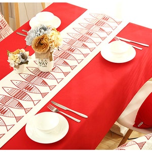May include: A red tablecloth with a white table runner featuring a repeating pattern of red fish. The table runner is decorated with a geometric design. The table is set with two white plates and a white bowl. There are two white cushions with red fish patterns on the chairs.