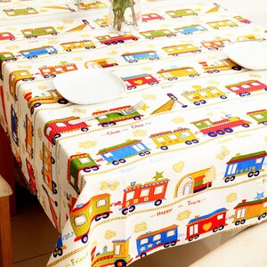 May include: White tablecloth with a colorful train pattern. The tablecloth features a variety of trains with different colors and designs, including red, blue, green, and yellow. The trains are all on a white background with a yellow rope design. The text "... Choo Choo...", "... Happy Train...", "... Friend...", and "... Little Train..." is printed on the tablecloth.