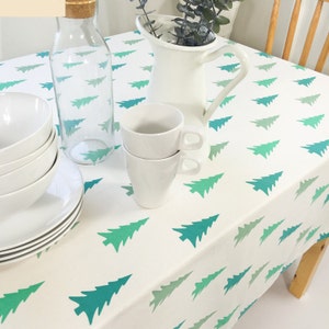 May include: White tablecloth with a repeating pattern of green ombre Christmas trees. The tablecloth is set with white dishes, a jug, and a glass bottle.