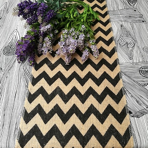 Può includere: Un runner da tavolo in juta con motivo a chevron nero e beige. Il runner è su una superficie con motivo bianco e nero con un mazzo di fiori viola.