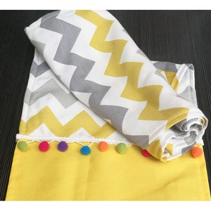 May include: A yellow and gray chevron patterned table runner with a white crocheted trim and colorful pom-poms.