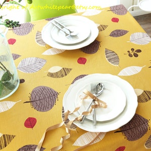 May include: A yellow tablecloth with a leaf pattern in shades of brown, white, and gray. The tablecloth has a subtle texture and is decorated with small red accents. Two white plates are set on the tablecloth, one with a gold ribbon and two forks.
