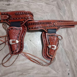 Può includere: Un set di cintura e fondina in pelle marrone. Il set presenta un'intricata lavorazione floreale, fibbie color argento e borchie decorative. La cintura ha una chiusura con fibbia e le fondine sono progettate per due pistole.