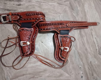 May include: A brown leather gun belt and holster set. The set includes a belt with a silver buckle, two holsters with silver buckles, and decorative tooling. The holsters have leather ties.