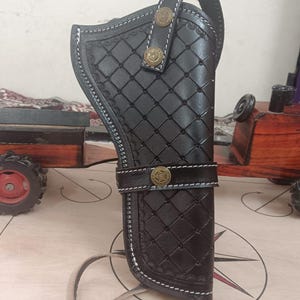 Puede incluir: Funda de cuero negro con un patrón de diamantes y costuras blancas. La funda presenta botones decorativos de color latón y una correa. Se adjunta un lazo para un cinturón. La funda está diseñada para guardar un arma de fuego.