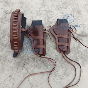 Può includere: Un set di fondine per pistole in pelle marrone con due fondine e una cintura con anelli per proiettili. Le fondine hanno una finitura marrone scuro e sono collegate da cinghie di cuoio. Anche la cintura è marrone.