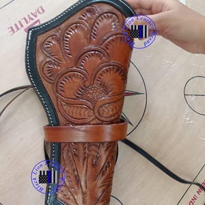 Pode incluir: Uma coldre de couro marrom com intrincados desenhos florais. Possui uma borda preta e uma al&ccedil;a. A imagem inclui o texto "Lion Leather Brand".