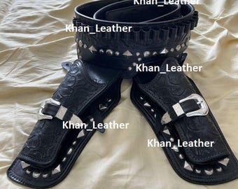 May include: Black leather gun holster set with silver-tone accents. The set includes a belt with bullet loops and two holsters. The holsters feature decorative embossed patterns and silver buckles. The text "Khan_Leather" is visible on the holsters and belt.