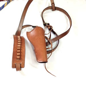 May include: A brown leather shoulder holster and ammunition holder. The holster is designed to be worn across the chest and back, with adjustable straps and a buckle closure. The ammunition holder has loops for holding bullets.