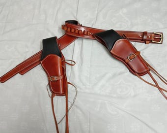 May include: A brown leather gun belt and holster set. The holsters are brown with black tops and secured with buckles. The belt has loops for bullets and a brass buckle. White stitching accents the edges of the leather.