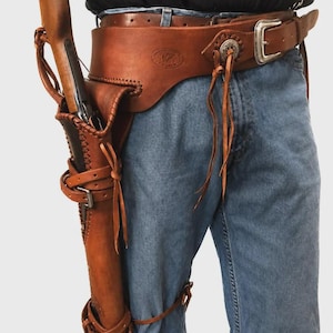 Op de afbeelding: Een bruine lederen wapengordel met een lange, verticale holster voor een geweer, vastgezet met meerdere lederen riemen en gespen. De riem is voorzien van een zilverkleurige metalen gesp en decoratieve lederen veterdetails.
