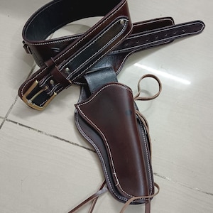 May include: A brown leather gun holster and belt set. The holster is designed to hold a handgun and is attached to a wide belt with a gold-coloured buckle. White stitching accents the edges of the holster and belt.