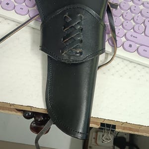 Puede incluir: Funda de cuero negro con una banda decorativa con cordones. La funda está diseñada para sujetar una pistola. El cuero está cosido a lo largo de los bordes y tiene una correa para asegurar el arma. La funda es probablemente para un usuario diestro.
