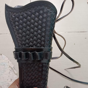 Könnte beinhalten: Schwarze Leder-Pistolentasche mit einem gewebten Korbgeflechtmuster. Die Tasche hat eine Schlaufe für einen Gürtel und einen Riemen. Die Tasche hat eine Reihe von Schlaufen für Kugeln. Die Tasche wird von einer Person gehalten.