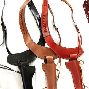 Op de afbeelding: Een verzameling leren pistoolholsters en schouderbanden in zwart, bruin en rood. De holsters zijn ontworpen om een ​​pistool te bevatten en de banden zijn verstelbaar. Het leer heeft zichtbare stiksels en een klassiek ontwerp.