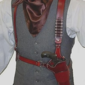 Op de afbeelding: Een rood leren schouderholster met witte stiksels, ontworpen om een revolver te dragen. Het holster wordt gedragen over een grijs vest en een wit overhemd, met een bruine zijden sjaal. Het holster heeft een riem en decoratieve kogelhouders.