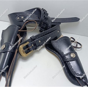May include: Black leather gun holster set with a belt and cartridge holder. The set includes two holsters, a belt with a gold-colored buckle, and a cartridge holder. The holsters have decorative silver-colored buttons.