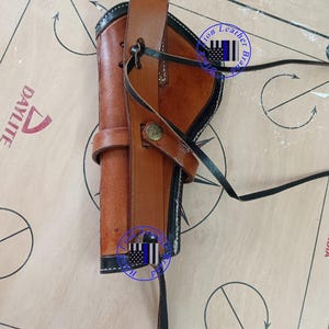 Pode incluir: Uma coldre de couro castanho com acabamento preto e uma correia. O coldre tem uma presilha para cinto e um fecho de bot&atilde;o. O fundo &eacute; uma superf&iacute;cie de cor clara com a palavra "DAYLITE" vis&iacute;vel. A imagem tamb&eacute;m cont&eacute;m uma marca d'&aacute;gua.