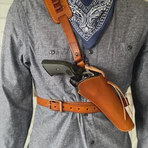 Könnte beinhalten: Ein Schulterholstersystem aus hellbraunem Leder mit integriertem Patronengurt, passendem Hüftgürtel und einem Holster, das einen schwarzen Revolver enthält. Die Ausrüstung wird über einem grauen Hemd und einem marineblauen Bandana mit weißem Muster getragen.