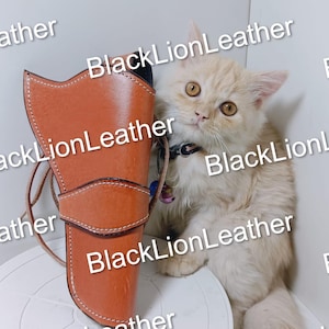 May include: A tan leather holster with visible stitching along the edges and a central strap closure. Beside the holster sits a fluffy, light-colored cat with large amber eyes and a dark collar featuring small, colorful hanging charms.