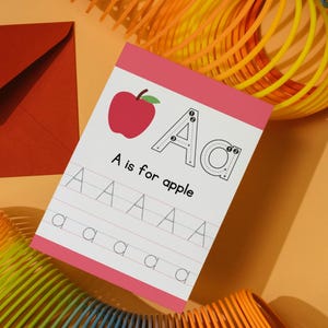 Peut inclure: Un bloc d'apprentissage avec une bordure rose présente la lettre "A" et une illustration de pomme rouge. Le texte "A is for apple" est affiché, ainsi que des guides de traçage pour la lettre "A". Une enveloppe rouge et un ressort coloré sont en arrière-plan.