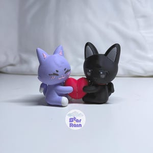 May include: Two cat figurines, one lavender and one black, holding a red heart. The lavender cat has a smiling face and white paws. The black cat has a red collar. The Star Rain logo is at the bottom.
