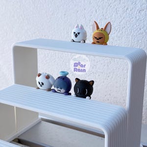 May include: A white two-tiered display stand with ribbed edges holding five small cartoon character figurines. The top shelf features a white creature and a brown-eared character. The bottom shelf holds a white bear, a purple figure, and a black cat.