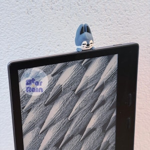 May include: A blue and white bunny-shaped accessory sits atop a black e-reader. The e-reader displays a grayscale image of textured lines. The accessory has a smiling face. The Star Rain logo is visible.