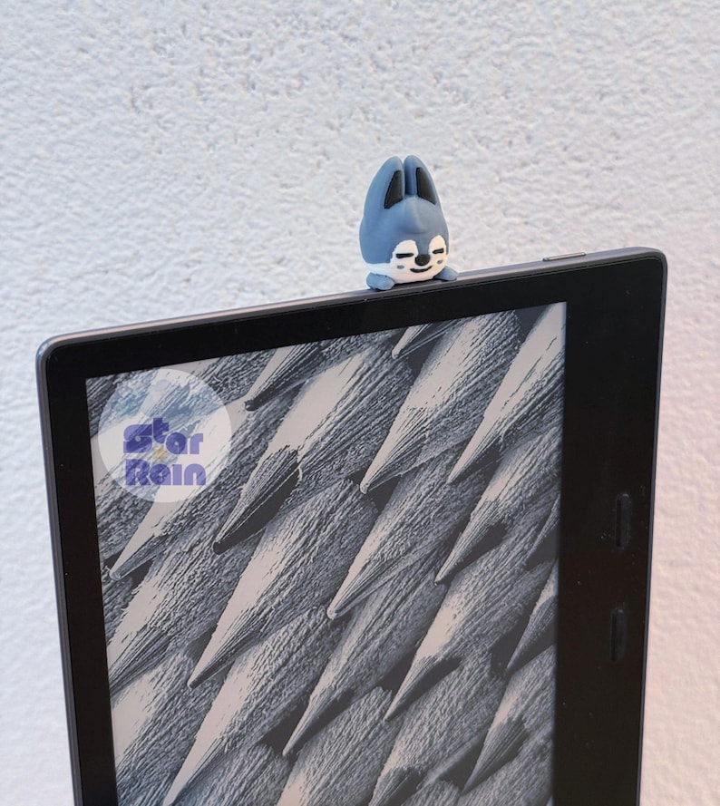 May include: A blue and white bunny-shaped accessory sits atop a black e-reader. The e-reader displays a grayscale image of textured lines. The accessory has a smiling face. The Star Rain logo is visible.