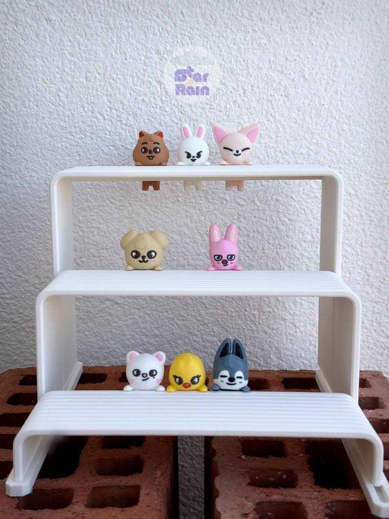 May include: A white, three-tiered shelf displays a collection of small, colorful animal figurines. The figurines include a bear, bunny, fox, dog, chick, and wolf. The shelf is set against a white brick wall.