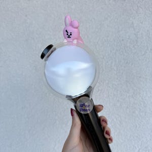 Op de afbeelding: Een zwart-witte lichtstaaf met een heldere bol en een roze konijnenfiguur erop. De handgreep is zwart en de bol is wit. Een kleine sticker met tekst is zichtbaar op de handgreep.