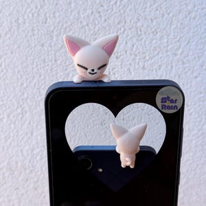 May include: A light pink fox-shaped phone charm with a smiling face and pink inner ears is perched on a black phone case. The phone case has a heart-shaped cutout. The text "Star Rain" is visible on the phone case.