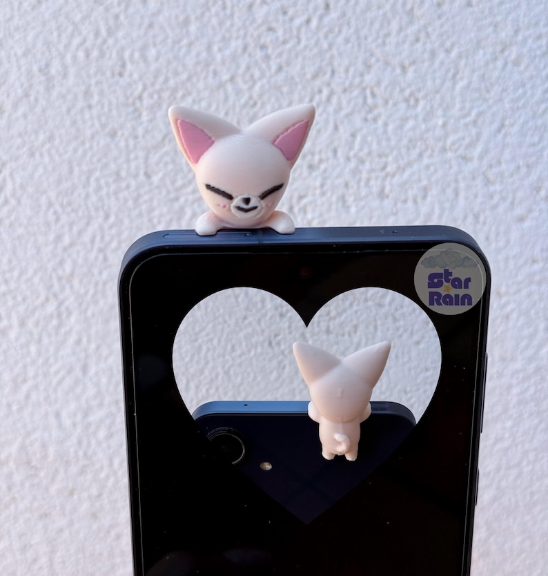 May include: A light pink fox-shaped phone charm with a smiling face and pink inner ears is perched on a black phone case. The phone case has a heart-shaped cutout. The text "Star Rain" is visible on the phone case.
