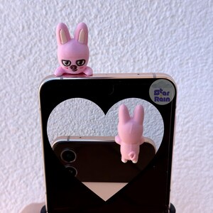 May include: A black smartphone with a heart-shaped cutout, featuring two pink bunny figures. One bunny is perched on top, and the other is visible through the cutout. The phone has a sticker that says "Star Rain".