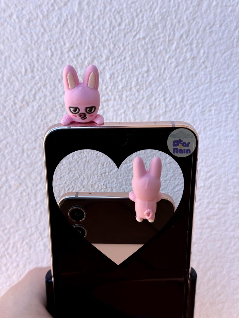 May include: A black smartphone with a heart-shaped cutout, featuring two pink bunny figures. One bunny is perched on top, and the other is visible through the cutout. The phone has a sticker that says "Star Rain".